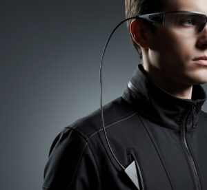 Close-up lateral de uma pessoa usando óculos inteligentes. Um cabo USB-C sai da haste do óculos e desce discretamente para dentro do bolso de uma jaqueta escura. Estilo fotografia de produto tecnológica.